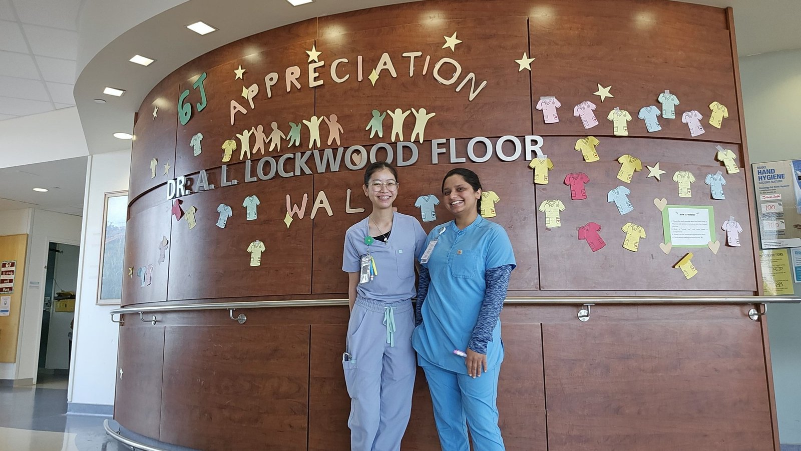 Two team members in scrubs in front of a staff appreciation wall